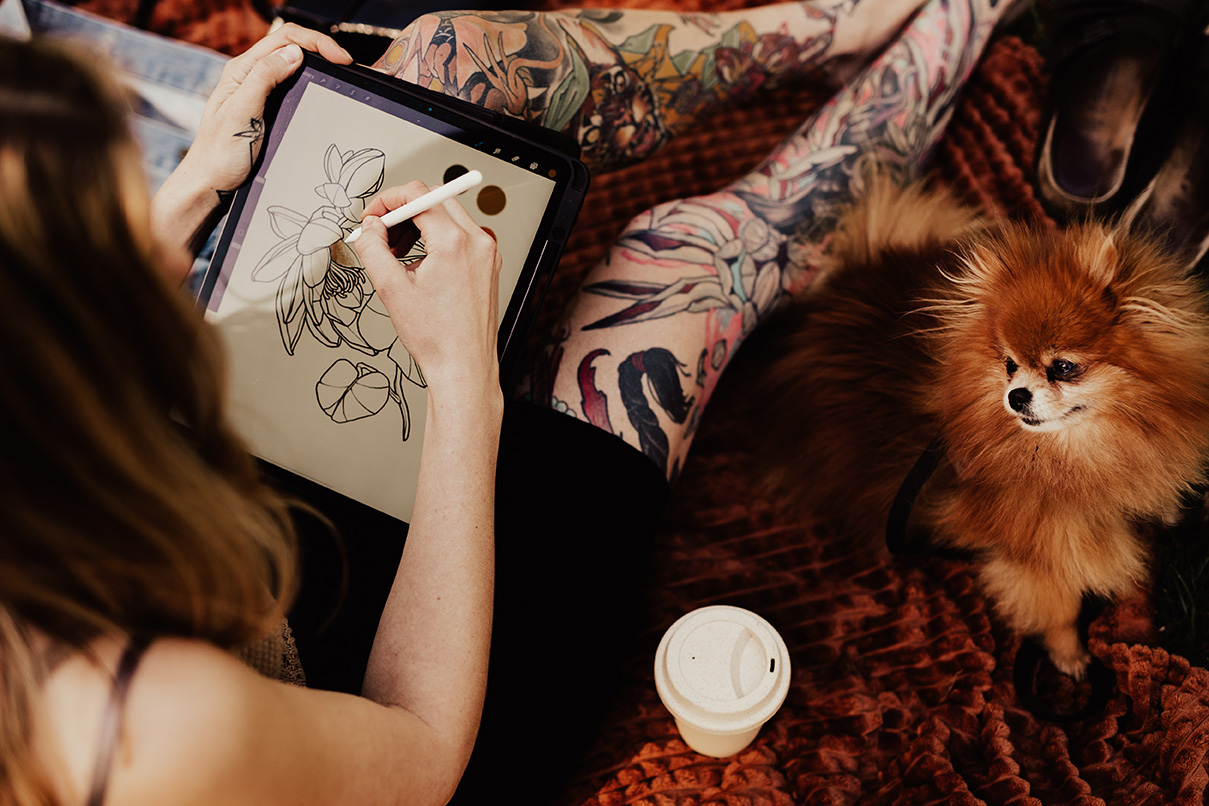 tatuerare sitter och ritar design på ipad med en pomeranian bredvid sig.